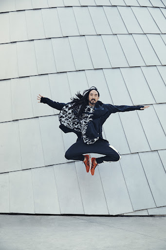 Full shot of long-haired adult male mid-air jump with arms outstretched, wearing athletic pants and jacket with printed scarf, smiling, against silver tiled wall.