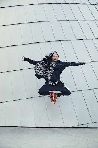 Full shot of long-haired adult male mid-air jump with arms outstretched, wearing athletic pants and jacket with printed scarf, smiling, against silver tiled wall.