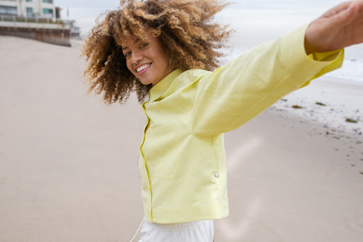Smiling woman with curly hair wearing a pale yellow button-down shirt spinning on a sandy beach