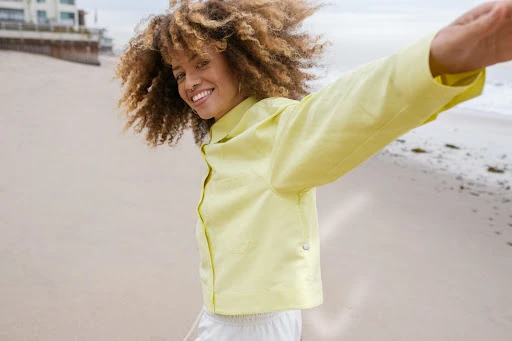 Smiling woman with curly hair wearing a pale yellow button-down shirt spinning on a sandy beach