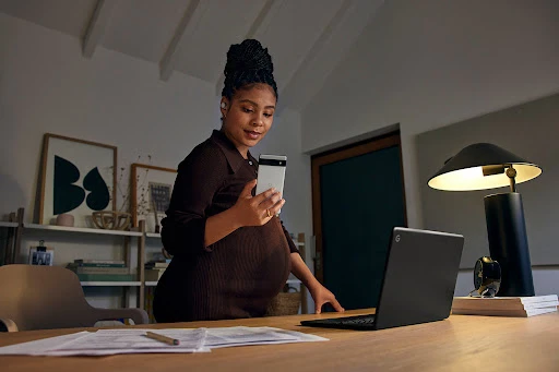Pregnant Black woman holding a smartphone standing at a wooden desk with a laptop and papers.