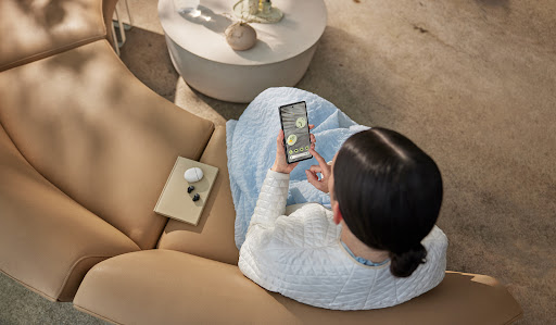 Woman uses phone on beige lounge chair next to wireless earbuds and a book, bathed in soft natural light in an indoor setting.