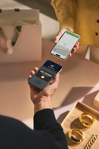 Two people exchange digital money via smartphones; shopping bag and jewelry tray visible; modern and contactless payment concept.