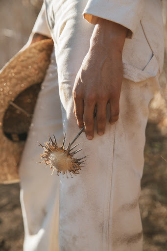 Partial view of a person holding a spiky burr, wearing a cream-colored outfit with a straw hat in an outdoor environment with natural light.