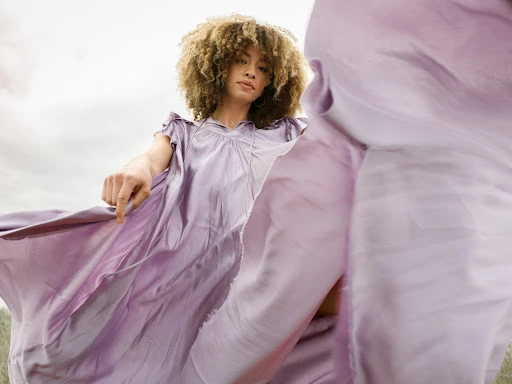 Woman in a flowing lilac dress posing outdoors with a dynamic fabric effect, soft light, and neutral background. Fashion and beauty shot.