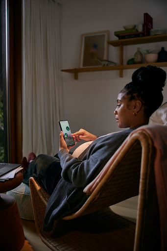 Pregnant adult female relaxes in wicker chair indoors using phone in softly lit home interior with neutral wall and dark wood window frame.