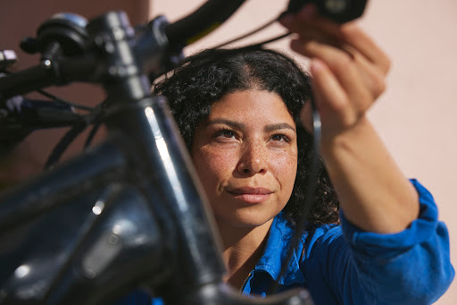 Close-up of a smiling woman in a blue shirt repairing the handlebars of a dark bicycle against a pale pink background in natural light.