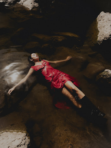 Woman in red dress and boots lies in a shallow river surrounded by rocks in an editorial fashion shot with soft lighting.