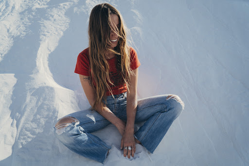 A woman smiles while seated cross-legged in ripped jeans and a red top in an outdoor setting with a bright, white background.