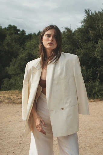 A stylish woman with long brown hair poses in a beige blazer and white pants against a backdrop of green trees and a cloudy sky.