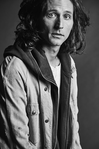 Black and white portrait of a man with long curly hair wearing a light denim jacket and dark hooded sweatshirt, looking at the camera.