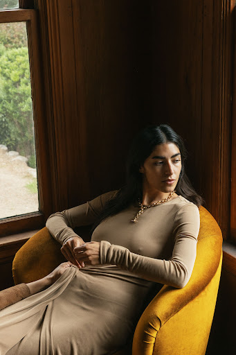 Woman in tan dress sits in yellow chair near window with natural light in an indoor setting, showcasing relaxed and elegant style.