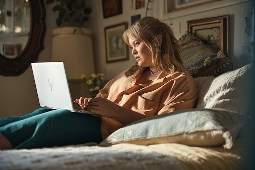 Young woman relaxes in bed using a laptop computer. The scene captures a cozy, personal moment in a warmly lit bedroom setting.