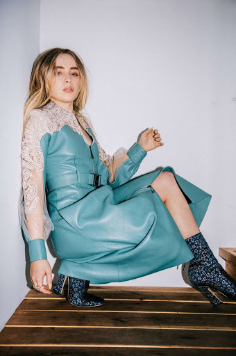 Blonde woman, Sabrina Carpenter, poses in a blue leather dress with lace sleeves and patterned boots against a white wall.