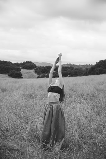 Monochrome image of a woman with arms raised in a field of tall grass against a backdrop of hills and sky.