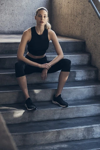 Fit woman sits on concrete steps in black athletic wear, looking at the camera with a neutral expression in an urban setting.