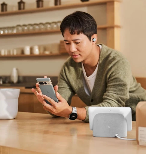 Man in a cardigan looking at a grey cell phone in a kitchen-like setting with a smart display on a wooden table and wireless earbuds.