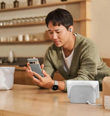 Man in a cardigan looking at a grey cell phone in a kitchen-like setting with a smart display on a wooden table and wireless earbuds.
