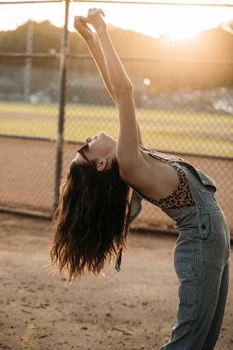 Woman in overalls arches her back in a sunlit outdoor scene. She is wearing sunglasses, with her arms raised and her head tilted back.