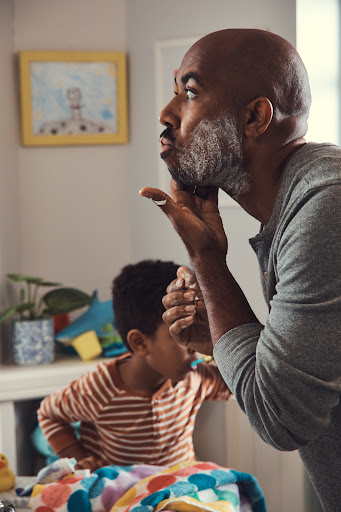 Black man applying shaving foam to his face in a bathroom with a young child brushing teeth behind him