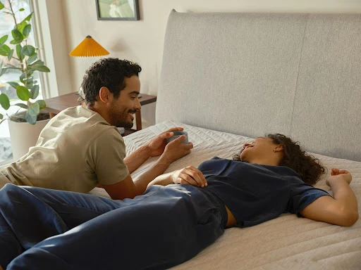 Couple relaxes on a bed with a drink in an indoor bedroom setting, featuring natural light and neutral colors.