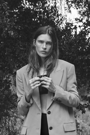 Monochrome portrait of a woman with long hair in a blazer, posed in front of a dense green leafy background in natural light.