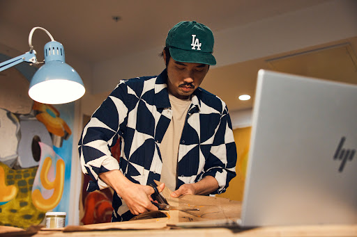 Adult male crafts indoors at a table with a laptop, wearing an LA baseball cap and patterned shirt, using scissors on a brown material.