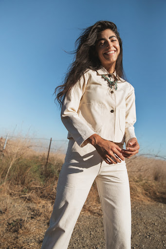 Smiling woman in a cream-colored suit stands outside on a sunny day, wearing a statement necklace against a blue sky background.