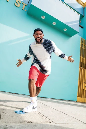 A cheerful adult male in a tie-dye shirt and red shorts strikes a playful pose on a city sidewalk, with a vibrant blue wall in the background.