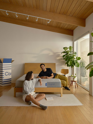 A man and woman relaxing in a bright bedroom, with the woman seated on a wooden floor and the man reclining on a bed.