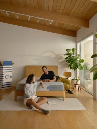 A man and woman relaxing in a bright bedroom, with the woman seated on a wooden floor and the man reclining on a bed.