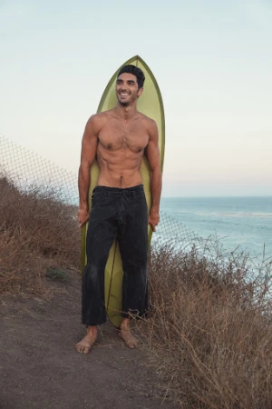 Shirtless man holding a surfboard standing on a coastal cliff path overlooking the ocean