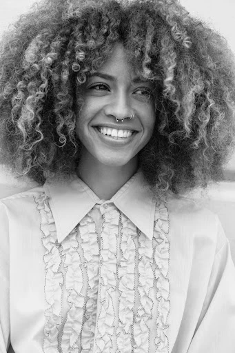 Smiling young woman with curly hair and ruffled shirt, in a close-up black and white portrait against a blurred light background.