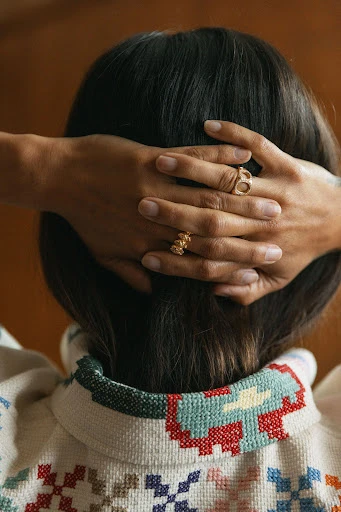 Brunette woman with hands behind head, wearing gold rings and a patterned jacket; close-up shot of the back of her head.