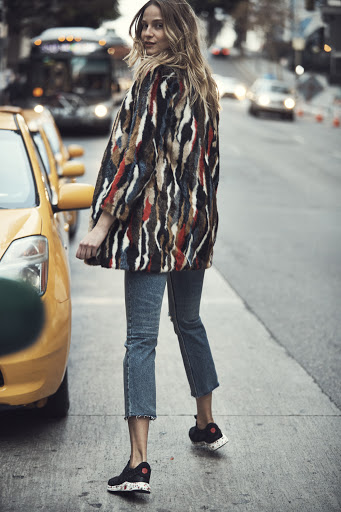 Adult woman in a colorful fur coat poses on a city street with a yellow taxi, showcasing a fashion forward look.