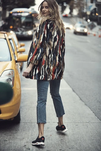 Adult woman in a colorful fur coat poses on a city street with a yellow taxi, showcasing a fashion forward look.