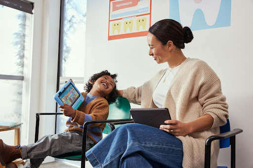 Child laughs while seated next to an adult in a waiting room, both holding tablet devices with educational dental posters in the background.