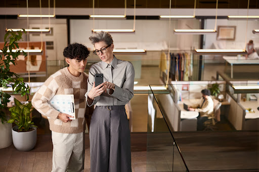Two professionals reviewing a mobile phone in a modern office setting with desks, plants, and bright overhead lighting.