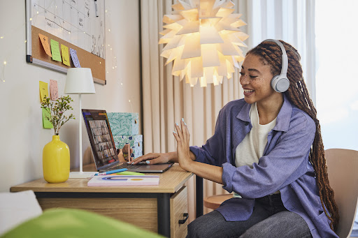 Smiling woman with braided hair and headphones is seated at a desk with a laptop, waving during a video call in an indoor setting.