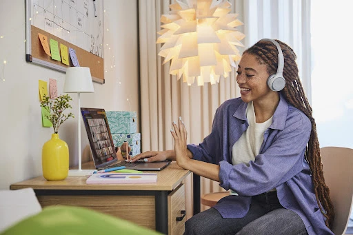 Smiling woman with braided hair and headphones is seated at a desk with a laptop, waving during a video call in an indoor setting.
