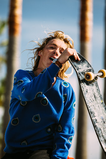 An eye-level shot of a young adult with long blond hair holding a skateboard while wearing a blue patterned sweater outdoors.