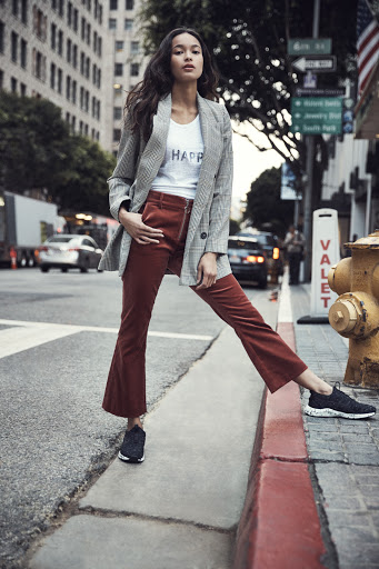 An adult female stands on a city street in a blazer and corduroy pants, posing next to a fire hydrant, with buildings and traffic in the background.