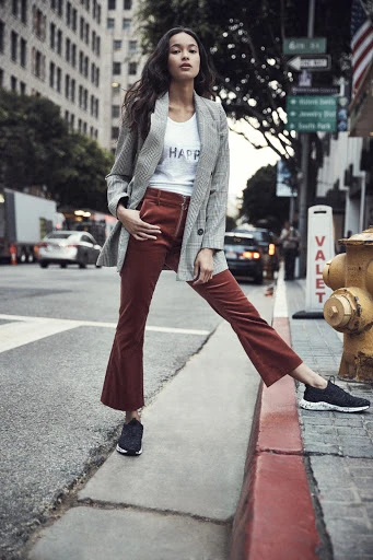 An adult female stands on a city street in a blazer and corduroy pants, posing next to a fire hydrant, with buildings and traffic in the background.