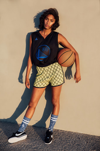 A model poses in athletic wear with a Golden State Warriors basketball jersey and a basketball in an outdoor setting.