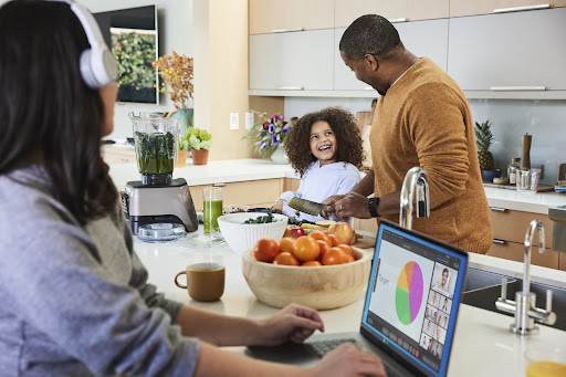 Family in modern kitchen: woman using laptop, child laughing with father preparing healthy meal with fruit, smoothie, and fresh vegetables.