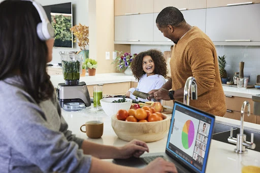 Family in modern kitchen: woman using laptop, child laughing with father preparing healthy meal with fruit, smoothie, and fresh vegetables.