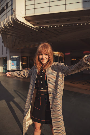 Woman with red hair wearing a houndstooth coat and dark skirt posing outside a building in Los Angeles.