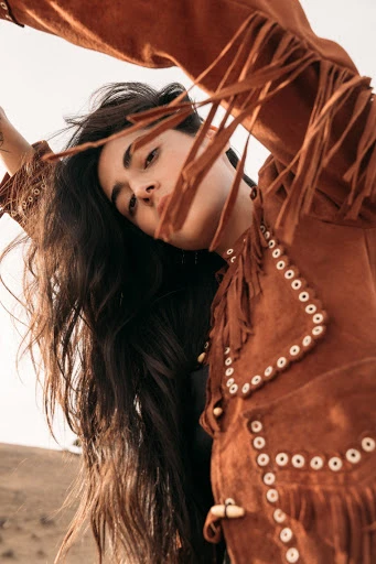 Brunette woman in a fringed suede jacket poses outdoors. She wears her long brown hair down and tilts her head looking at the camera.