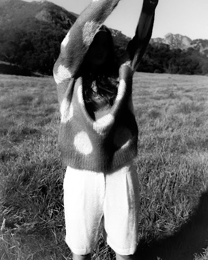 Black and white outdoor shot of a person with arms raised wearing a polka dot sweater, white shorts, and mountains in the background.