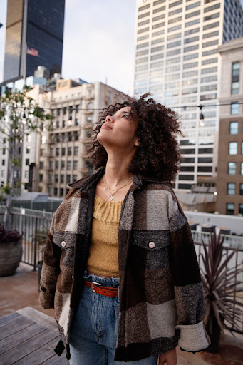 Adult female with curly hair and denim jeans stands on a rooftop in an urban setting with tall city buildings in the background.